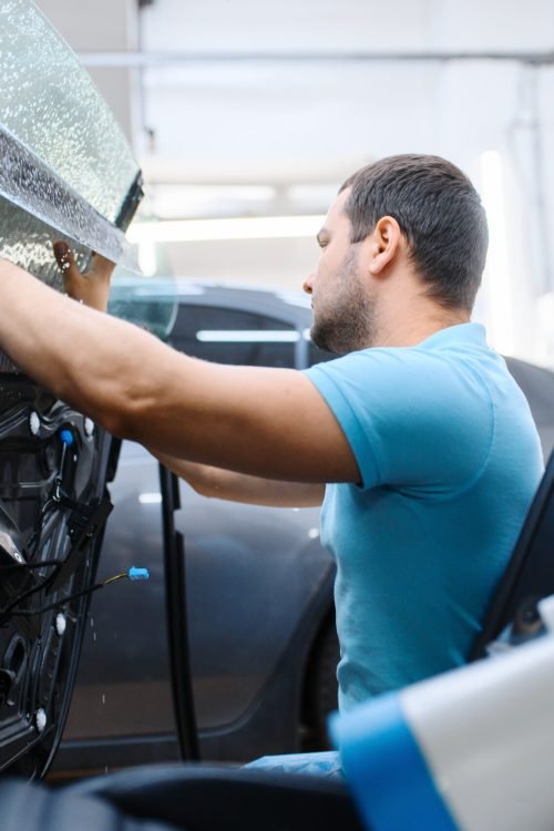 Male specialist installs wetted car tinting, tuning service. Mechanic applying vinyl tint on vehicle window in garage, tinted automobile glass